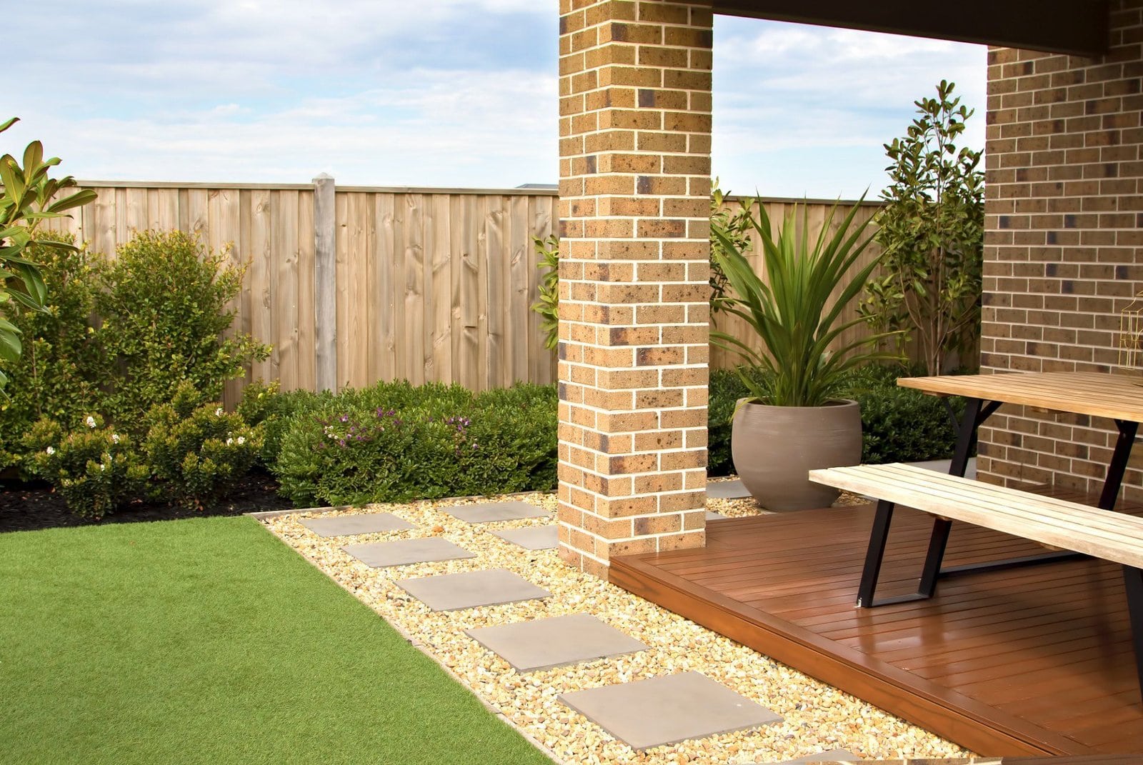 A modern backyard with a wooden deck featuring a picnic table. The deck is adjacent to a brick pillar and bordered by neatly trimmed greenery. The yard, enhanced with turf products, has a stone path leading through a well-maintained lawn, and a wooden fence encloses the area.