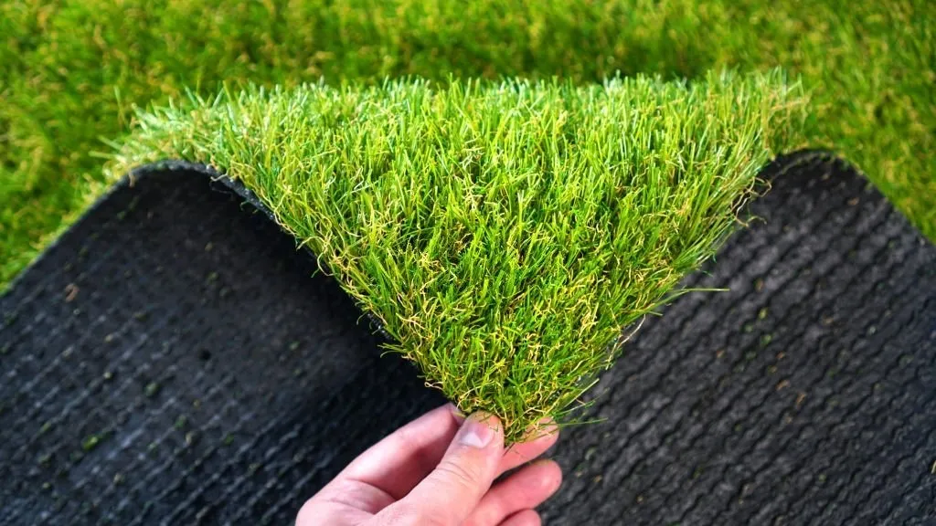 A close-up image of a hand lifting a corner of a square piece of artificial grass, revealing the black rubber backing underneath. This vibrant green pet turf contrasts sharply with the dark backing, perfect for enhancing outdoor spaces in Paradise Valley AZ.