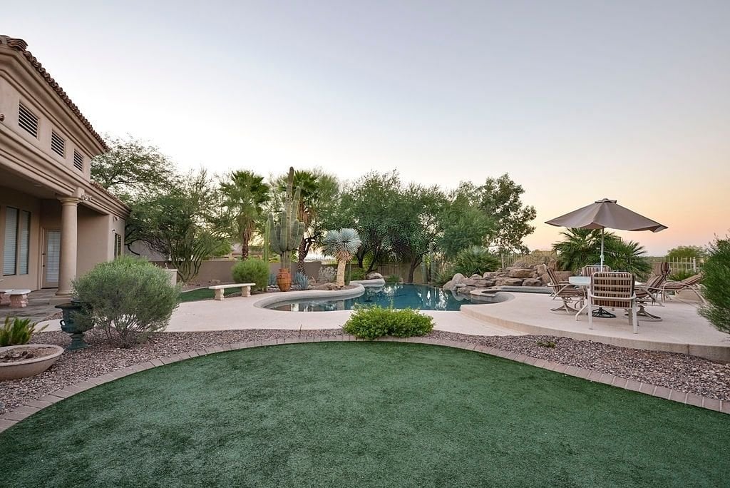 Spacious backyard featuring a lush green lawn, enhanced by Scottsdale artificial turf, a large pool with a spa, and an elegant patio area. Poolside, there are lounge chairs under a large umbrella for relaxation. The yard is landscaped with desert plants and trees, offering a serene oasis-like atmosphere.