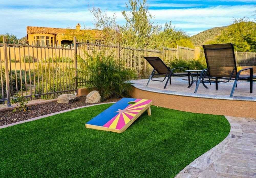 A backyard scene in Scottsdale, AZ, with a cornhole game board on artificial turf in the foreground, two lounge chairs on a paved patio with a curved edge, and desert vegetation. An adobe-style house and a hill with a mix of greenery are in the background under a blue sky.