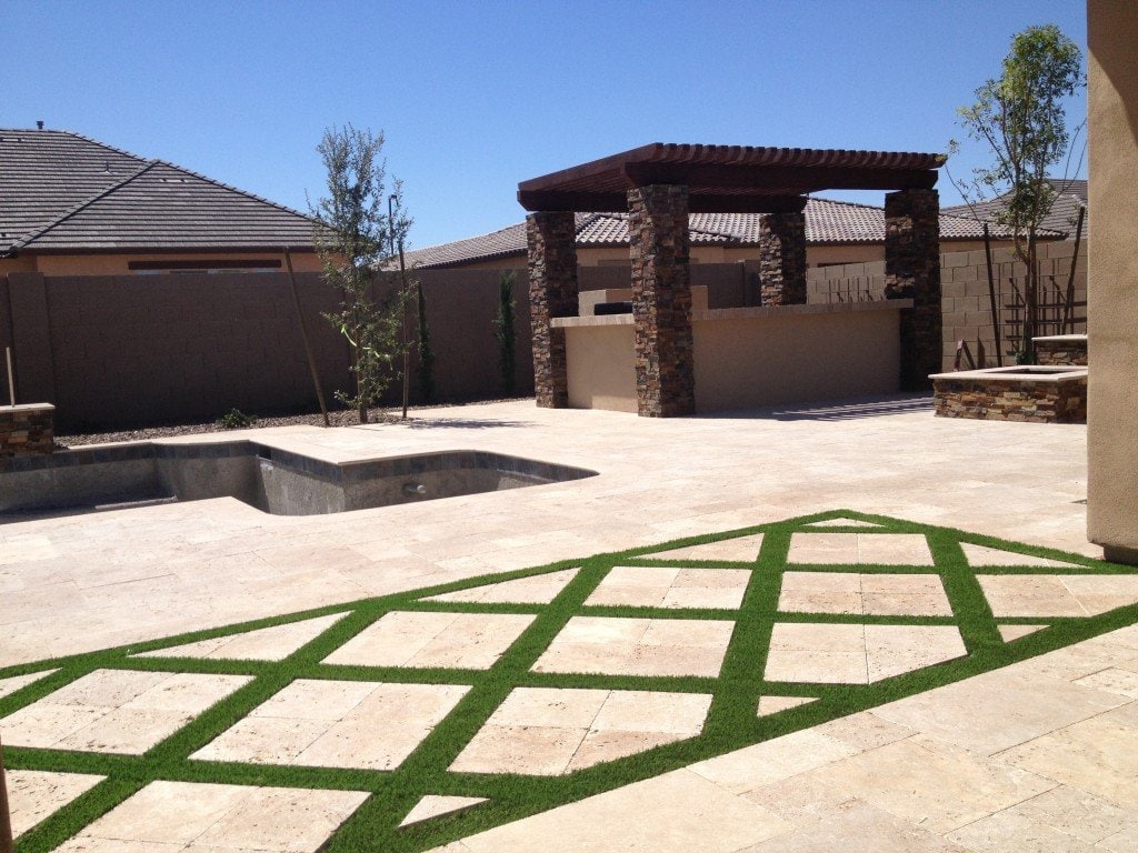 A backyard in Paradise Valley with a large paved patio featuring green grass strips arranged in a diamond pattern. The yard includes an unfinished pool in the foreground and a covered outdoor kitchen area with stone columns and a wooden pergola. Reach out for a free quote from professional artificial grass installers.
