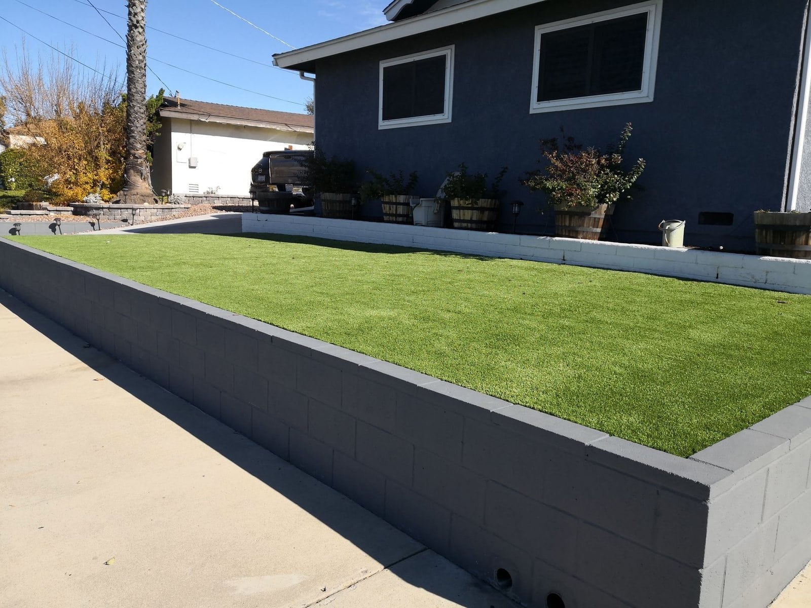 A well-maintained raised garden bed with artificial turf is situated in a residential backyard in Tempe, AZ. The bed is bordered by a low grey brick wall, and several potted plants line the edge near the house. A driveway and parked vehicle are visible in the background, showcasing quality work by Scottsdale Artificial Turf.