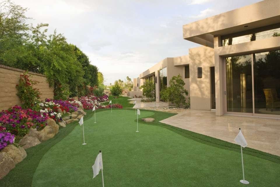 A modern house in Scottsdale, AZ boasts a neatly manicured yard featuring pet-friendly turf and a putting green. The green has several flags and is surrounded by colorful flowers, trimmed bushes, rocks, and well-maintained landscaping. A large glass door opens to the backyard.
