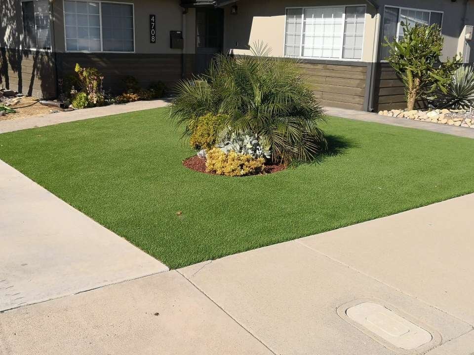 A well-maintained lawn in front of a house with neatly trimmed grass, expertly crafted by artificial grass installers. Surrounded by a concrete walkway and driveway, it features a small landscaped area with various green plants and decorative rocks, offering homeowners in Paradise Valley a glimpse of tranquility. Contact us for a free quote!